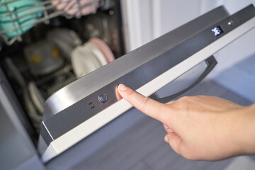 Female hand pressing start button on dishwasher door with clean dishes inside modern kitchen.