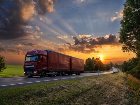 Red delivery truck driving on the asphalt road in rural landscape at sunset