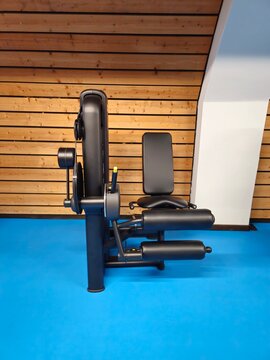 A sleek black leg extension and curl machine stands on a blue gym floor against a modern wooden wall.