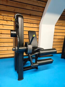 A sleek black leg extension and curl machine stands on a blue gym floor against a modern wooden wall.