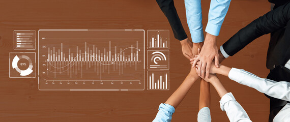 A diverse business team collaborates around a table with hands joined over digital data analysis and financial statistics on a modern office desk. Trope