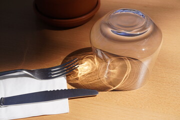 Round outdoor café table set with cutlery on napkins, an upside-down glass, and a potted plant as decoration. Sunlight creates soft shadows, giving the scene a cozy and inviting atmosphere