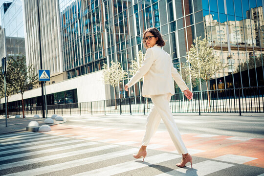 Confident young businesswoman in white suit walking in a sunny urban city environment with modern glass buildings