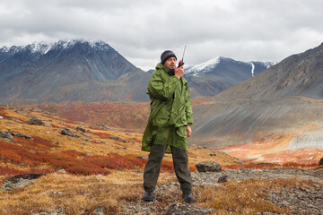 A man, European, 50 years old, stands with a walkie-talkie in his hands against a backdrop of mountain peaks. He's talking on the radio.