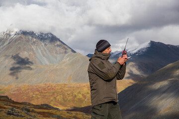 A man, European, 50 years old, stands with a walkie-talkie in his hands against a backdrop of mountain peaks. He's talking on the radio.