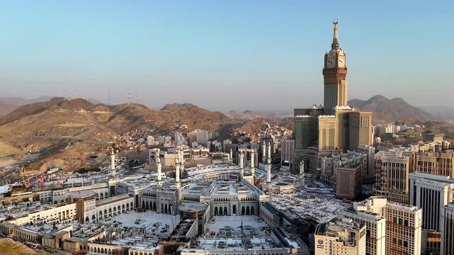 drone footage of Mecca, Saudi Arabia, filmed at sunset. The video captures the vast urban landscape with residential buildings, modern towers, and surrounding mountains bathed in golden hour light