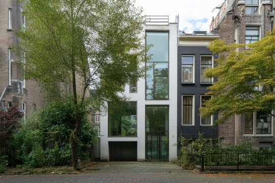 Modern architectural design stands prominently among traditional buildings on a quiet street in Amsterdam surrounded by greenery and trees during a clear day