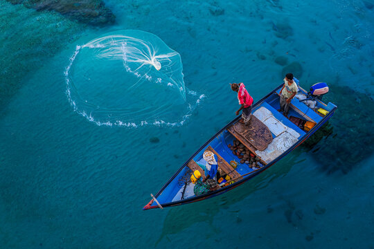 Fishermen on wooden boat casting net in crystal-clear blue water, aerial view of traditional fishing