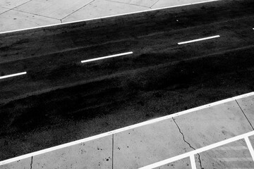 A black and white image of a deserted paved road featuring white lane markings and cracked concrete curbs, symbolizing direction, journey, and urban solitude.