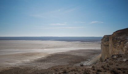 A panoramic view of the Boszhira Valley on the Ustyurt Plateau in Kazakhstan. The rugged terrain with rocky cliffs meets the vast salt flats, extending to the horizon under a deep blue sky.