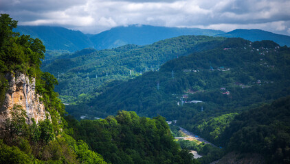 a striking view of a tree-covered cliff with distinct stratified rock formations overlooking a lush valley with a winding road, set against a backdrop of misty mountains and a dynamic sky