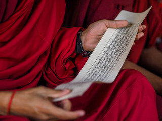 Close-up of monk in red robes reading sacred text or prayer on traditional paper during ritual or study