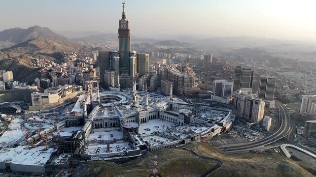drone footage of Mecca, Saudi Arabia, filmed at sunset. The video captures the vast urban landscape with residential buildings, modern towers, and surrounding mountains bathed in golden hour light