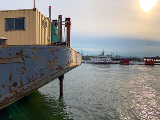 Weathered barge with a small cabin, moored on calm harbor water. In the background, cranes, ships and port infrastructure fade into hazy sunset sky with soft sun flare. Maritime/industrial scene