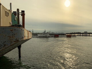 Weathered barge with a small cabin, moored on calm harbor water. In the background, cranes, ships and port infrastructure fade into hazy golden-hour sky with soft sun flare. Maritime/industrial scene
