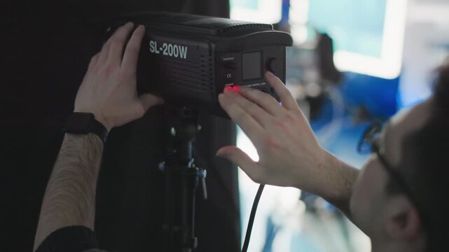 Technician adjusting power button and brightness dial on SL-200W LED studio light mounted on tripod bracket amid blue-lit studio backdrop during broadcast production setup with cables visible