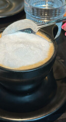 Frothy cappuccino in black cup with spoon on saucer - coffee shop, café, restaurant concept