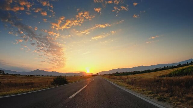 POV driving on straight road across fields, sunset light, clouds