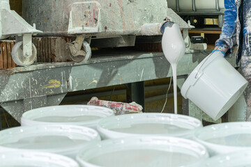 Manual paint pouring process in industrial workshop. White pigment flowing from industrial spout into containers during manual paint production in factory.