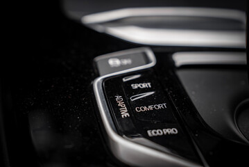 Close up view of the BMW X4 drive mode selector panel with SPORT, COMFORT, ECO PRO, and ADAPTIVE options on a glossy black surface with silver accents. © Aerial Film Studio