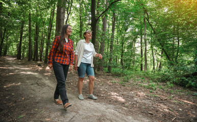 Fototapeta premium Two women walking together in a green forest, smiling and enjoying nature on a summer day. Friendship, leisure, outdoor activity, and healthy lifestyle.