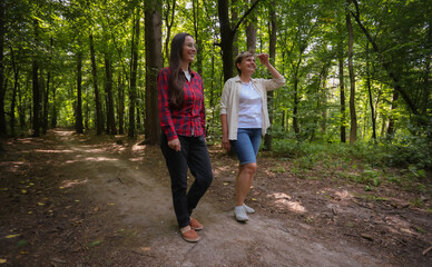 Obraz premium Two women walking in a forest, smiling and enjoying a sunny day surrounded by tall trees. Friendship, leisure, outdoor lifestyle, and nature exploration.