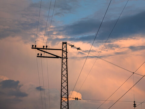 Silhouette of a lattice steel electrical power pylon and transmission wires against a dramatic evening sky of soft pink, orange, and deep blue clouds during sunset or twilight.