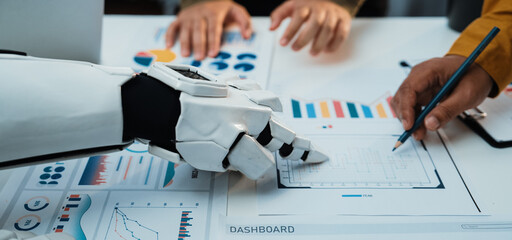 A human and a robotic hand collaborate over various data visualizations and reports, highlighting modern technology's role in shaping analytical processes in business.Noogenesis