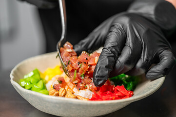 Chef in black gloves preparing poke bowl with raw tuna, rice, vegetables, and peanuts. Close-up of colorful, fresh ingredients in modern healthy cuisine