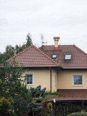 Cozy, traditional residential house in muted yellow with a textured terracotta tile roof, complete with skylights, a brick chimney, and a small antenna array, surrounded by a lush, varied green garden