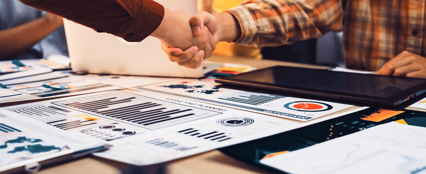 A diverse business team celebrates a successful partnership with a handshake over various financial reports and data analytics in a bright modern office environment. SACTR