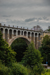 Obraz premium Majestic stone viaduct with multiple arches standing tall over a lush green valley, showcasing historical engineering and architecture against a dramatic cloudy sky in late summer season.