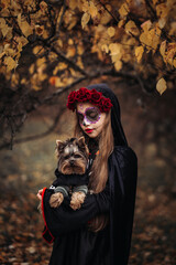 A young woman in a Day of the Dead costume: purple and violet skull makeup, red roses on a wreath, and a black cloak. She held a small dog against the backdrop of an autumn park with fallen leaves.