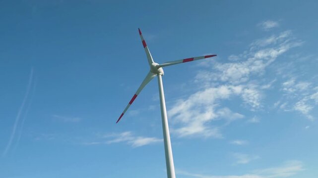A tall white wind turbine stands against a bright blue sky with wispy clouds