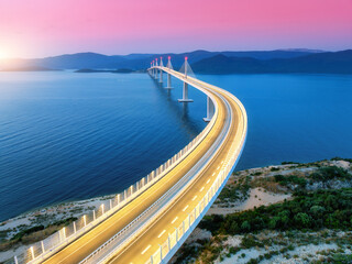 Obraz premium Aerial view of modern Peljesac bridge and blue sea at sunset. Croatia. Bridge with illumination, mountains, reflection and pink sky at sunset. Top drone panoramic view of road, street lights at dusk