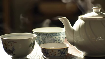 A collection of decorative porcelain teacups and a teapot, steaming gently in the light. - Powered by Adobe