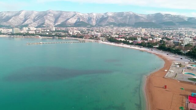 Central beach of Gelendzhik in snow. Aerial view. Zia at resort. Caucasus Mountains in snow. In Gelendzhik Bay you can see pier of sea station. People walking on snowy beach.