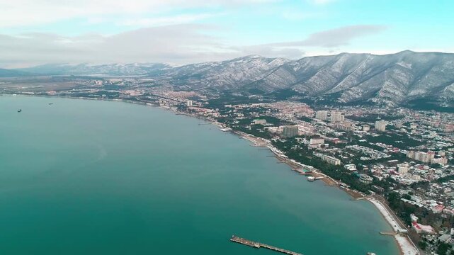 Central beach of Gelendzhik in snow. Aerial view. Zia at resort. Caucasus Mountains in snow. In Gelendzhik Bay you can see pier of sea station. People walking on snowy beach.