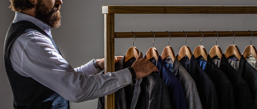 Handsome bearded fashion man in classical costume suit. Male suits hanging in a row. Men clothing, boutiques. Man suit, tailor in his. Tailor, tailoring