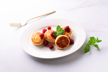 Cottage cheese pancakes with honey, raspberry and mint leaves on white ceramic plate on white marble table close up. Healthy homemade dessert.