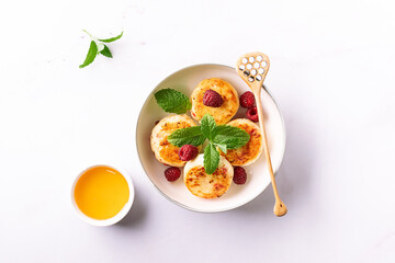  Cottage cheese pancakes with honey, raspberry and mint leaves on white ceramic plate on white marble table close up. Healthy homemade dessert.