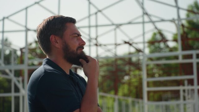 young man touching beard thoughtfully while looking into distance beside metal frame structure rural setting sunlight casting soft shadows on face and shirt