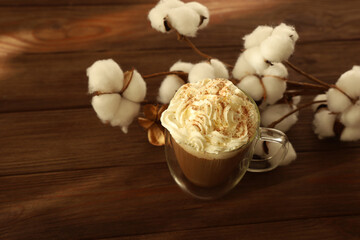 Warm coffee topped with whipped cream and spices beside soft cotton on a wooden table