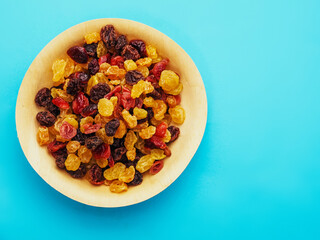 A bowl of dried fruit with yellow and red raisins, cranberries. The bowl is on a blue table surface. Natural high sugar and glucose content. Popular snack.