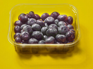 A plastic tray of blueberries sits on a bold yellow color background. The tray is clear and filled with fresh organic fruits ready for consumption. Healthy diet concept. Natural product.