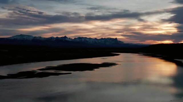 River flows towards snowcapped mountains under a dramatic sunset sky