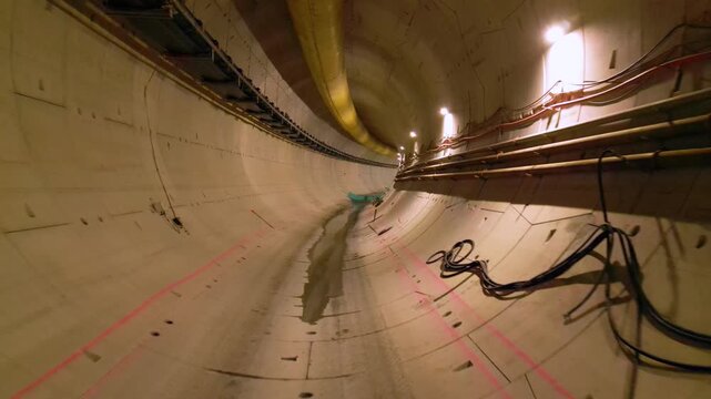 Long empty tunnel view showing modern reinforced concrete lining with visible seams, utility installations, service pipelines and faint lighting along curving underground transportation passageway

