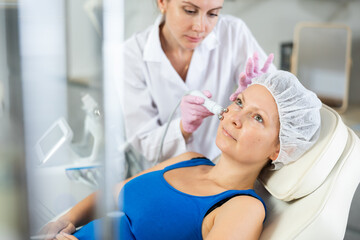 Fototapeta premium Young doctor doing RF lifting procedure for face lifting of adult woman laying in modern spa center. Smoothes wrinkles and tightens skin machine