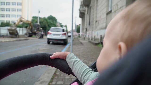 A baby with short hair in a green sweater sits in a stroller and watches attentively and intently the work of a construction excavator on the other side of the road on a cloudy summer day......