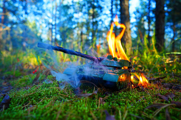 Naklejka premium Military Toy Tank Catches Fire in Forest During Bright Sunny. Smoke rises against of tall trees. Game of War. Battle tank during burning. War costs, military spending. Battle toy tank in burnt.